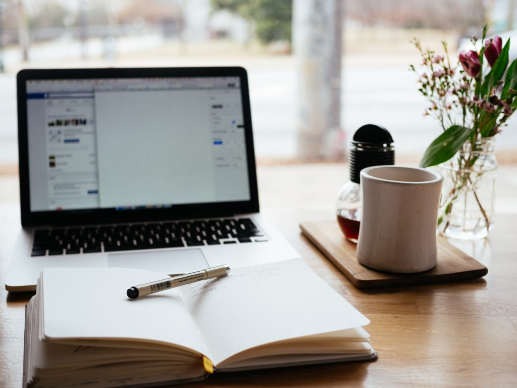 An image of an Apple mac, beside a cup and an open notebook with pen in it.